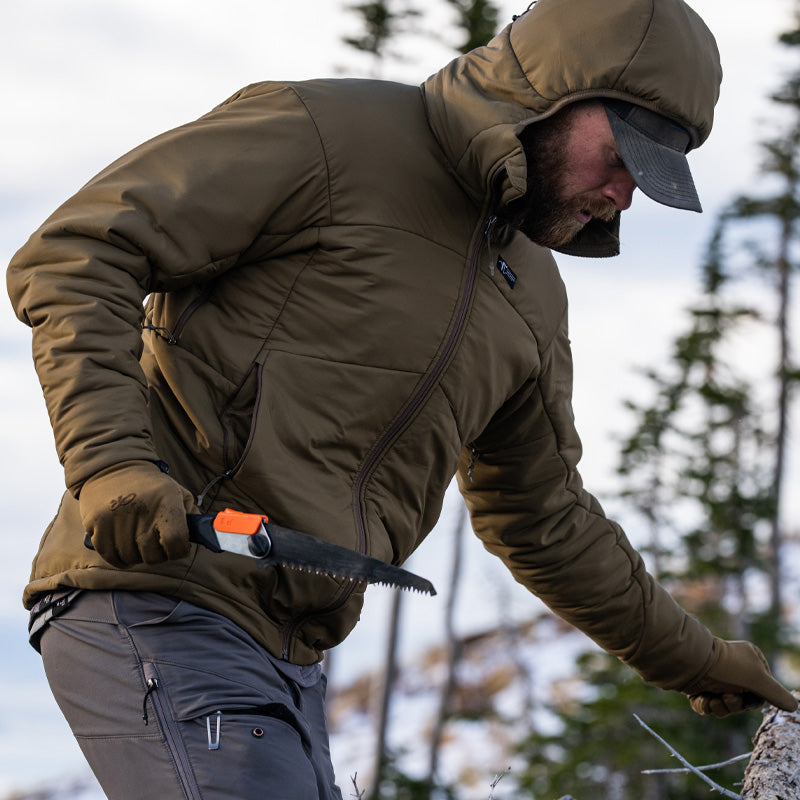 A man traversing a mountain side while holding a hand saw and wearing the Stone Glacier Cirque Jacket in muskeg.