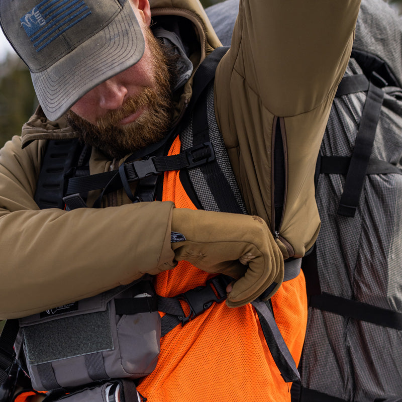 A backcountry hunter unzipping the heat vents found on the Stone Glacier Cirque Jacket while wearing a backpack and binocular harness.