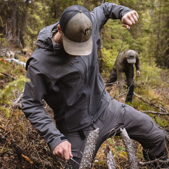 Two backpackers setting up camp and cutting cordage while wearing the Stone Glacier De Havilland Jacket in granite grey.