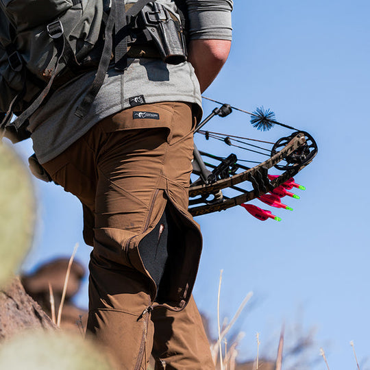 A close-up image of an archery hunter with bow in hand, hiking over a ridge line while wearing the Stone Glacier De Havilland LITE Pants in muskeg.