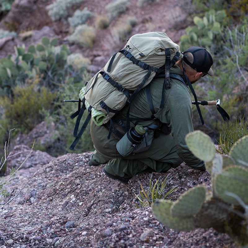 A backpacker descending through rocky terrain with a Stone Glacier Hydro Holster installed and a Nalgene bottle positioned within.